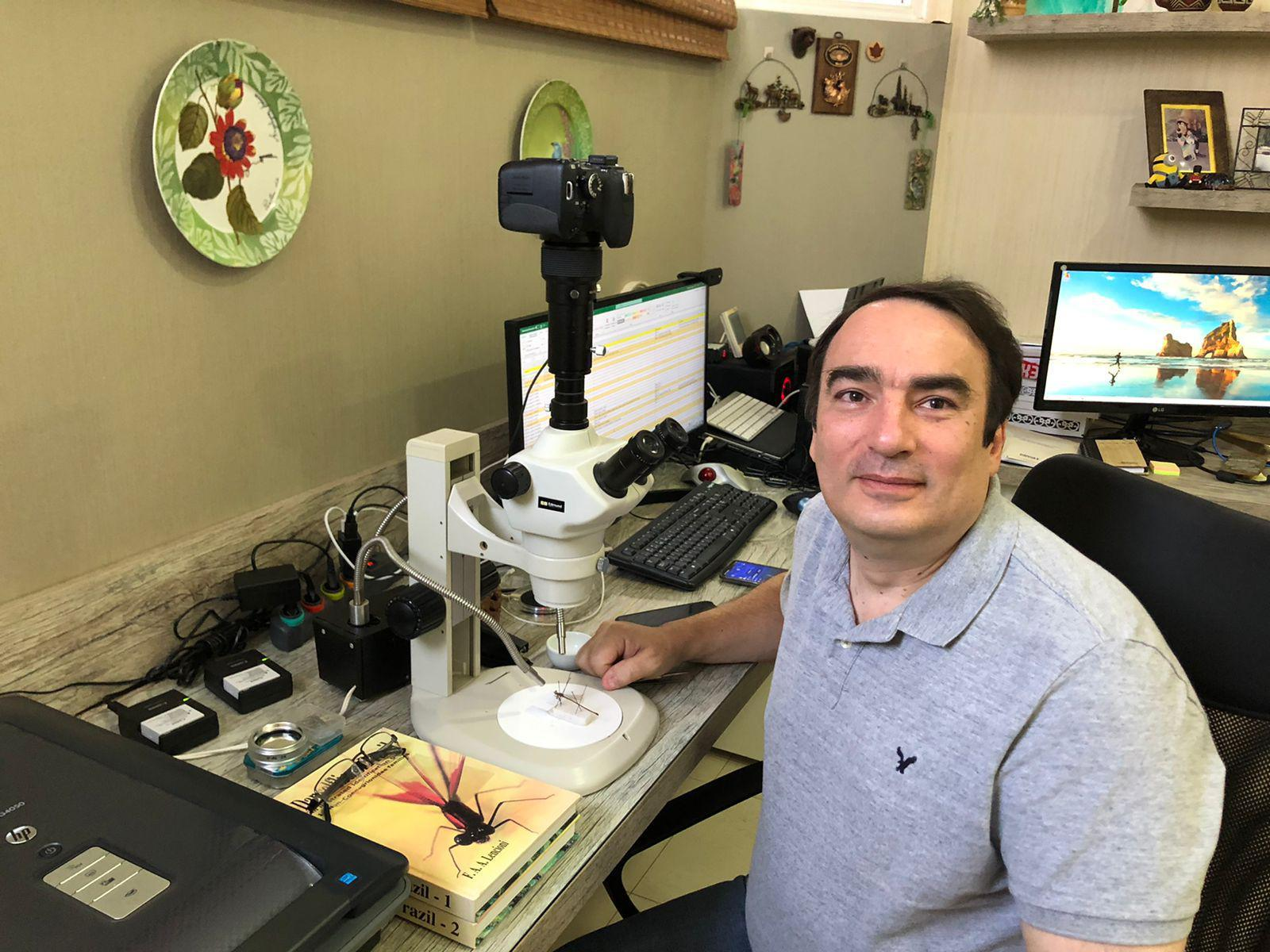 Frederico A. A. Lencioni em seu espaço de trabalho, com estereomicroscópio e material de estudo.